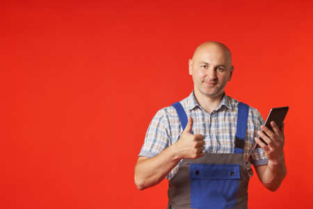 Bald man in a work suit holds a phone with one hand, and with the other shows a raised finger gesture, a photo on a red background. The client evaluates the work of the builders.の写真素材