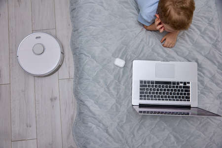 Little blond boy in white wireless headphones uses a modern laptop on the bed, gets new knowledge...