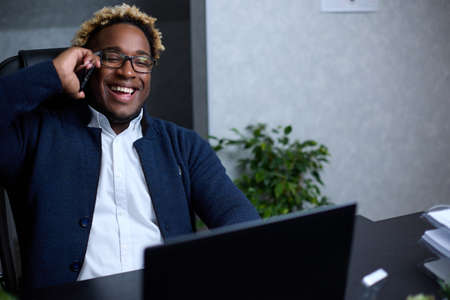 Smiling friendly African man talking on phone in office, having pleasant conversation with client, customer, receiving good news or offer, marketing manager making business call,chatting with friendの写真素材