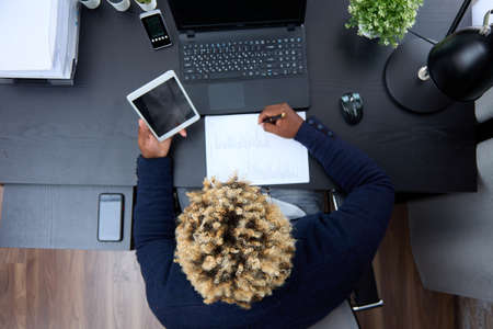 Top view of an African-American employee working with a laptop and tablet, sitting at an office...