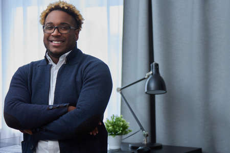 Confident African business leader or manager, leaning against his desk in the office, arms crossed...