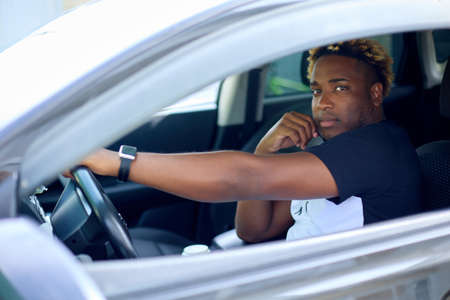 Confident, serious African-American man in a T-shirt is sitting in his car. A black man with an African hairstyle looks at the camera through the side window of a vehicleの写真素材