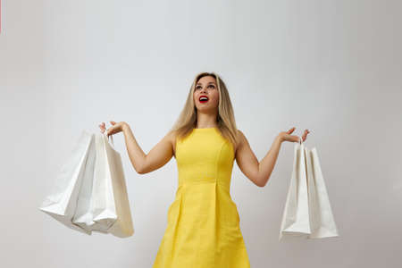 Young attractive blonde in a yellow summer dress with shopping bags in her hands opened her mouth with delight and looks up. Picture of the concept of purchase, discounts and salesの写真素材