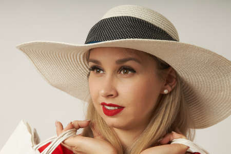 Close-up of a beautiful young blonde in a red dress and a straw hat posing with shopping bags on a white background. An attractive customer with a sweet smileの写真素材