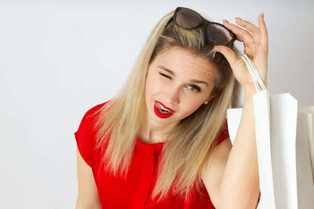 Close-up of a beautiful woman taking off her sunglasses and winking in a red dress. A girl holds shopping bags isolated on a white background.の写真素材