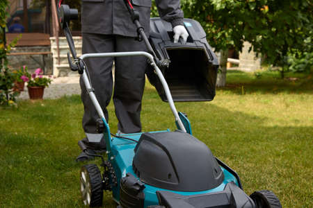 Man has cleaned the lawn mower basket and is ready to get back to work. A worker in overalls and...