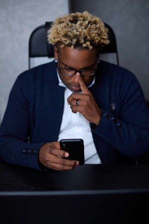 Worried male freelancer looking at the smartphone screen while working in office received bad news, received a message about fraud. Thoughtful African man with a worried expression uses a mobile phoneの写真素材