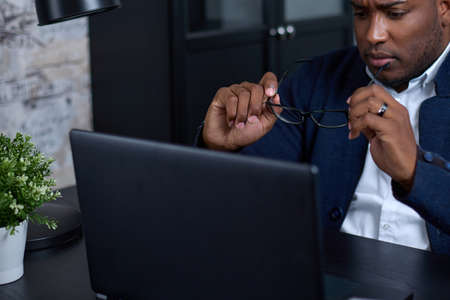 Tired African-American employee in formal clothes, took off his glasses, holds glasses in his hands,...