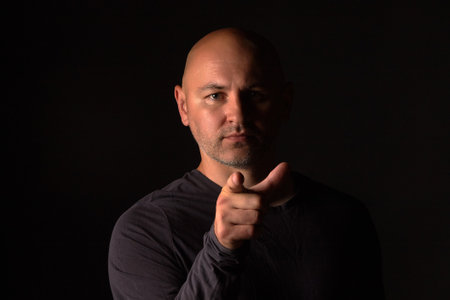 Serious bald man stands on a black background and points his finger at the camera. Portrait of a sporty man in a dark turtleneck with a shadow on half of his faceの写真素材