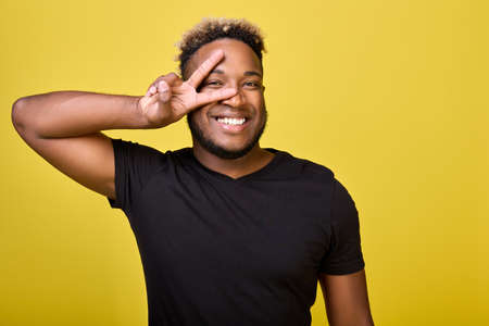 The symbol of peace shows with his right hand a dark-skinned guy in modern everyday clothes on a yellow background. Smiling broadly African-American demonstrates peace and kindness around himの写真素材