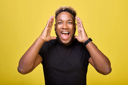 A black fan in a black T-shirt and smartwatch shouts loudly in the stadium for his favorite team. A joyful fan of a football fan shouts from the stands with his mouth wide open.の写真素材