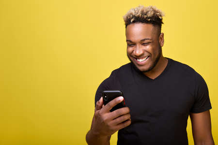 A curly joyful man reads the news on his phone. A dark-skinned strong young man smiles looking at the screen of a smartphone, on a yellow background in a black T-shirtの写真素材