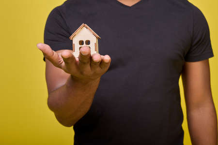 Photo of torso of black guy without head in frame in black everyday T-shirt, in his hand small toy house. Small house made of wooden construction set on palm of African-American on yellow backgroundの写真素材