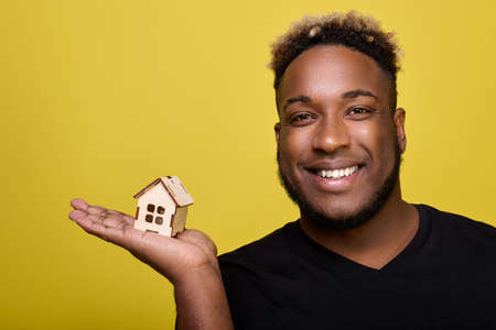A smiling black guy holds a small light toy house in the palm of his hand, advertises a real estate agency. Curly African-American demonstrates a house construction set in his handの写真素材