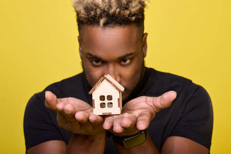 Serious black guy stares intently at small house holding in the palm of his hand in front of him. Close up of toy house in the hands of African-American who looks it intently with a serious lookの写真素材