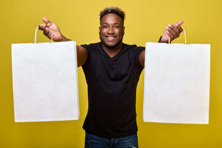 A handsome black guy smiling, stretched his arms forward, holding two white paper bags of shopping from the store. Smiling guy demonstrates his bargains made on the days of big discountsの写真素材