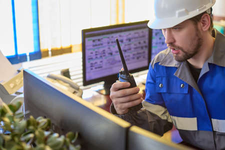 Serious Caucasian unshaven worker in overalls and helmet uses a walkie - talkie sitting in the control room . Automated workplace of a dispatcher or operator of a modern enterpriseの写真素材