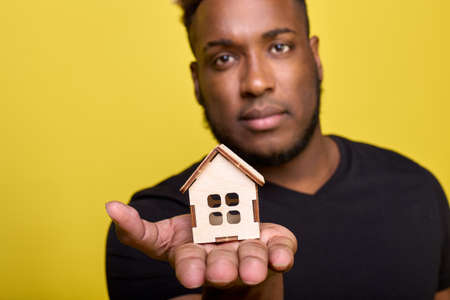 A dark-skinned cute guy stretches a toy house in the palm of his hand as a symbol of warmth and comfort. Calm peaceful man shows a small wooden house as a symbol of the future of your own big houseの写真素材