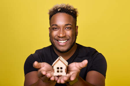 A small paper house with a window and a roof on the palms of a smiling black guy. In the hands of a cheerful African-American a small house assembled from a wooden construction setの写真素材