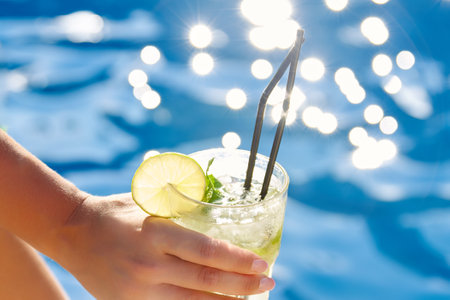 Close up full glass cool mahitto with ice in hand tanned girl against background blue sparkling water. Against background of pool with clear water, girl with cocktail in hand is resting on sunny dayの写真素材