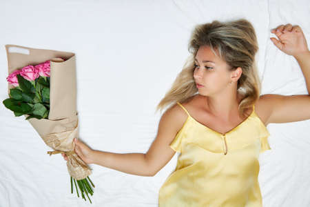 Young girl in silk yellow peignoir lies on bed and holds in her hand fragrant bouquet of white pink roses. Huge bouquet of romantic roses in the hand of girl who is relaxing in hotel room on seaの写真素材