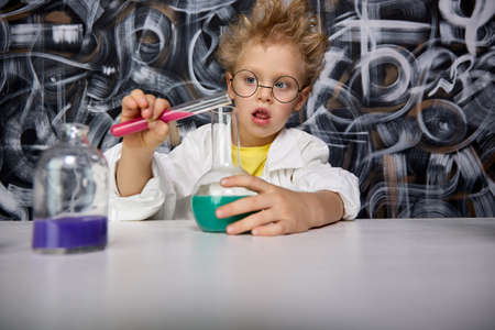 Beautiful little boy scientist is intrigued and excited, exploring colored chemical liquids in the laboratory using test tubes of different sizes. Cute scientist with glasses mixes reagents in the labの写真素材