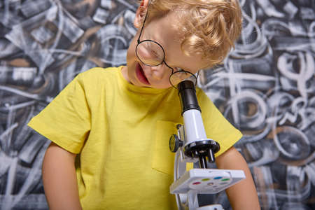 Closing one eye boy stares intently at eyepiece of microscope through magnifying glass stage table on which crystals are placed. Child is fond of science and studies different substances in microscopeの写真素材