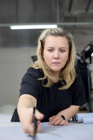 Vertical photo of a dressmaker cutting a piece of fabric with professional scissors in a sewing workshop. Making a piece of clothing or upholstery for furniture made of fabric in a studio workshopの写真素材