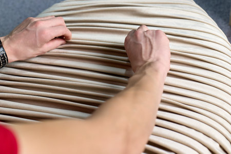 Man pleats fabric to order at a furniture factory. Process of creating folds or draping beige fabric by hand on a furniture pouf.の写真素材