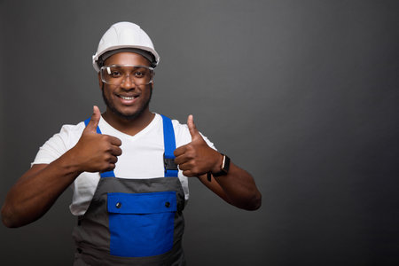 Smiling, dark-skinned worker in a gray helmet and construction jumpsuit with a blue pocket shows his...