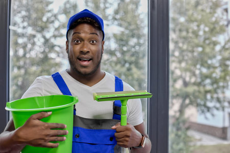 Surprised black cleaner holds a green bucket and a brush for washing windows and floors. An employee...