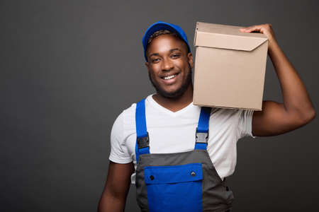 Against gray background, strong black guy confidently holds cardboard box with office supplies on one shoulder. A cheerful loader easily carries weights in a box on his shoulder holding only one handの写真素材