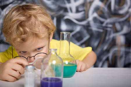 Blonde boy in glasses carefully examines through magnifying glass blue liquid in glass tube. Chemical substance in flask aroused genuine interest in the young scientist in the childrens laboratoryの写真素材
