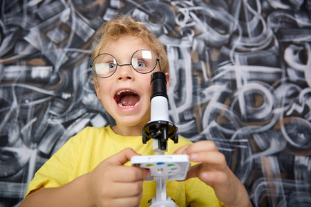 The boy screams with delight the field of what he saw on stage through microscope. An enthusiastic child in yellow T-shirt and round glasses rejoices at successful experiment in childrens play lab.の写真素材