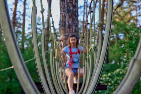 Woman is preparing to walk along a swinging suspended walkway in protective gear. Rope road on the background of a blurred climber in a rope park high above the groundの写真素材