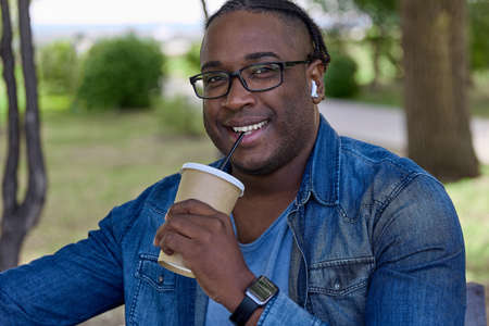 Young black man with glasses rests in the park on a weekend drinking hot chocolate from a paper cup with a straw. On a sunny summer day, a handsome African man relaxes in a city parkの写真素材