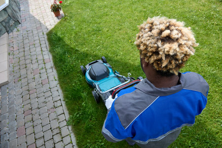 Top view of an African man in overalls mowing green grass in a modern garden with a lawn mower. A black man with an Afro hairstyle uses a lawn mower in the backyard. Professional lawn care service.の写真素材