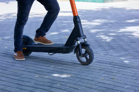 Close up of a man getting ready to ride an electric scooter in a city park. A man in brown sneakers and jeans in a park, with one foot steps on the foot platform of an electric scooterの写真素材