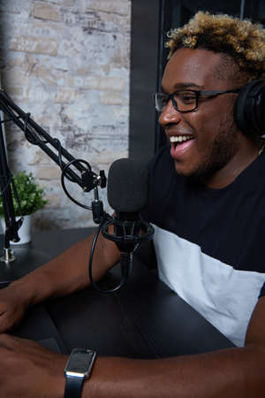 Joyful black radio presenter in a modern loft-style studio hosts the program. A young guy listens to an online lecture, uses a laptop and headphones to communicate with the lecturer and classmatesの写真素材