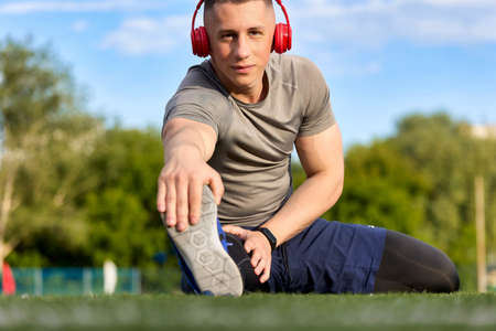 Serious and focused athlete prepares for the game, listening to his favorite music in headphones, trains at the stadium. A handsome young man in sportswear stretches, warming up on the streetの写真素材