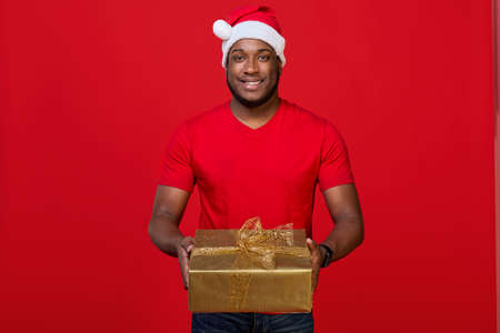 Portrait of a cheerful black guy with a toothy smile in a Santa hat and a red T-shirt giving a Christmas gift in a golden package with two hands on a bright red backgroundの写真素材