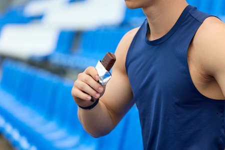 Man in a blue sports jersey rests in the stand for fans, eating a chocolate bar. A muscular athlete snacks on a protein bar while resting after a workout at the stadiumの写真素材