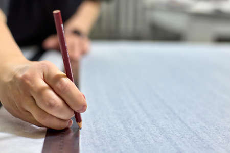 Close-up of the seamstress hands make a drawing on the fabric using a ruler, a template and a pencil. Professional seamstress making pattern with pencil. Process of cutting and sewing.の写真素材