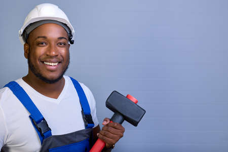 Attractive black man in a white helmet and work clothes looks at the camera and smiles while holding...