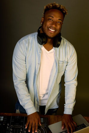 An unshaven guy with dark skin smiles while at the DJ console and a laptop on a gray background. Stylish dj mixing music on professional controller in booth at nightclub partyの写真素材