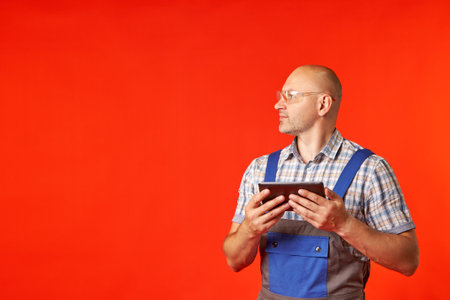 Bald man in work clothes and protective glasses holds a phone in his hands and looks in the other...