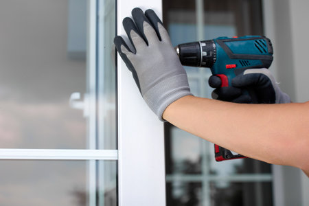 Close-up of a workers gloved hand holding an automatic screwdriver repairing a plastic door. An experienced master maintains plastic windows and doors with professional toolsの写真素材