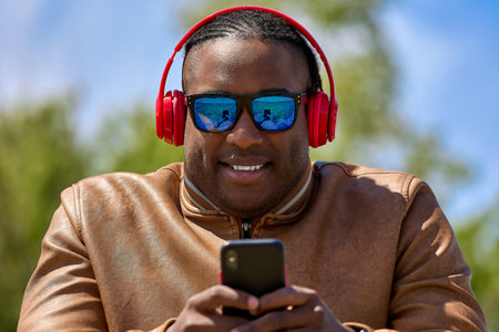 Dark-skinned man in sunglasses is relaxing in park, playing in casino online on a smartphone. African guy in wireless headphones of red color plays on the Internet on phone resting on the streetの写真素材