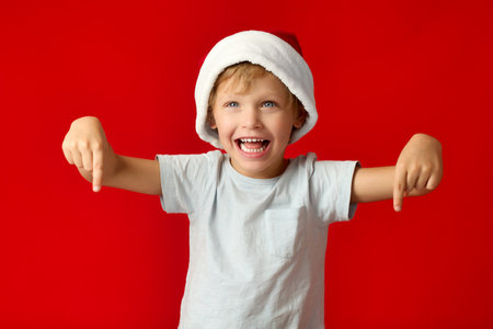 Cheerful little boy in funny gnome hat points his fingers down on red background. Funny boy smiles broadly and points his fingers downの写真素材
