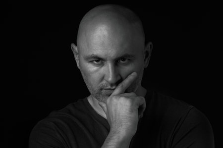 Bald man holds his hand on his chin and looks seriously at the camera, a darkened photo on a black background. Immersion in a state of sadness, darkness, despairの写真素材